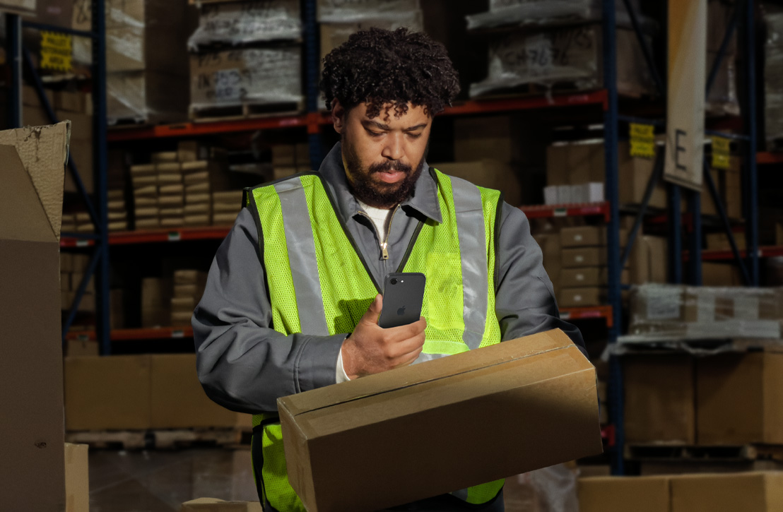 iPhone, black, held by a person in a warehouse, wearing a high-visibility vest, checking the screen while holding a cardboard box