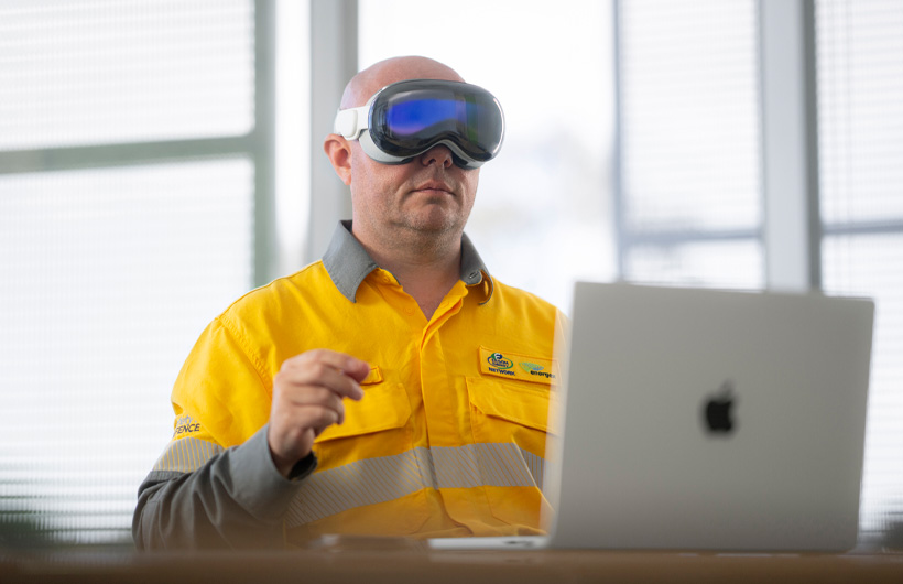 En ansatt med Apple Vision Pro på hodet gjør en knipebevegelse med hendene, mens en MacBook Pro i sølv står åpen på skrivebordet