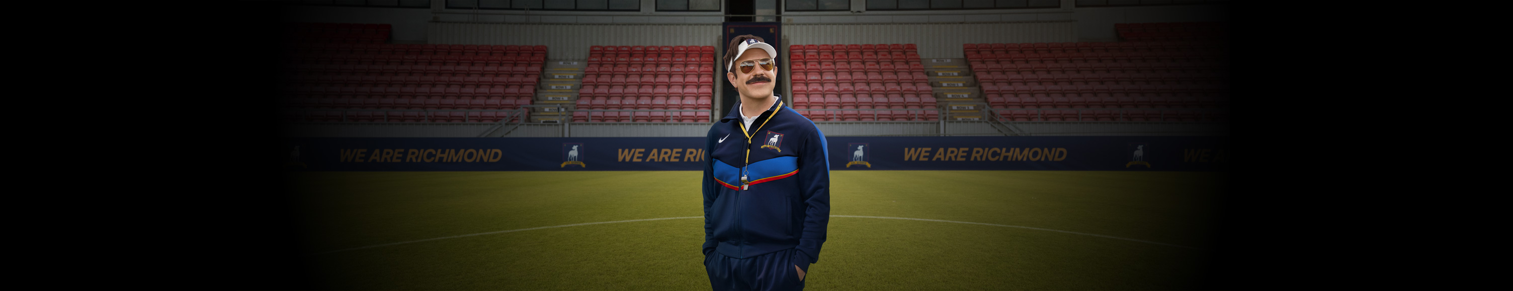 Promoci&oacute;n de Apple TV, Jason Sudeikis aparece sonriendo en el campo del AFC Richmond, vestido con su uniforme de entrenador, gafas de aviador y una visera.